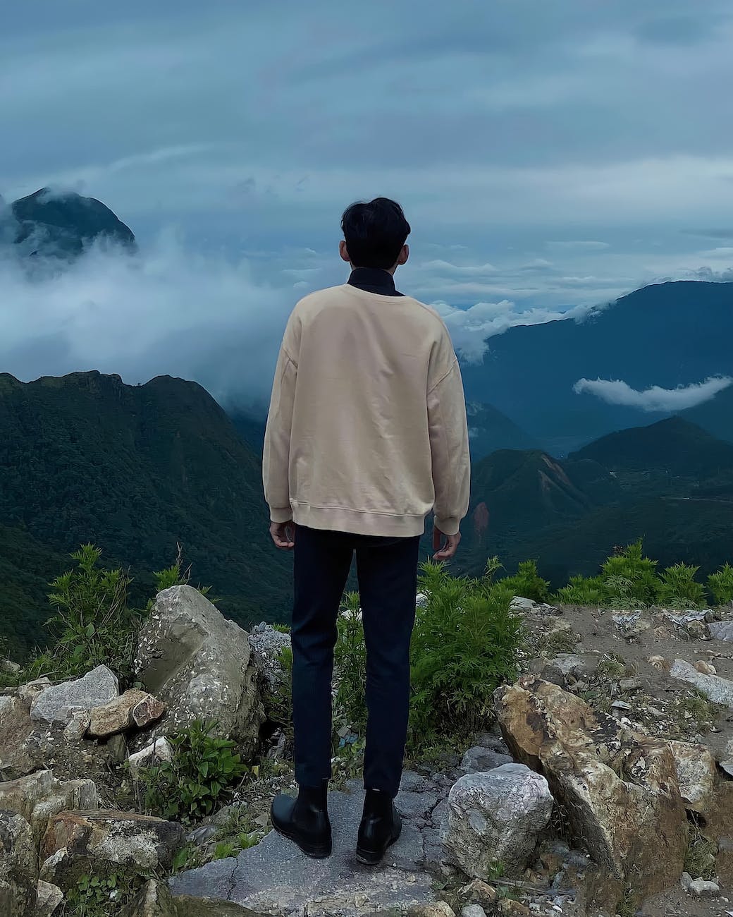 man in white hoodie standing on rock formation looking at mountains